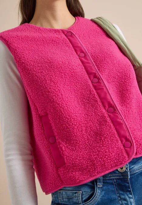 Frau trägt pinke Teddyweste über weißem Langarmshirt und Jeans, mit grünem Taschengurt.