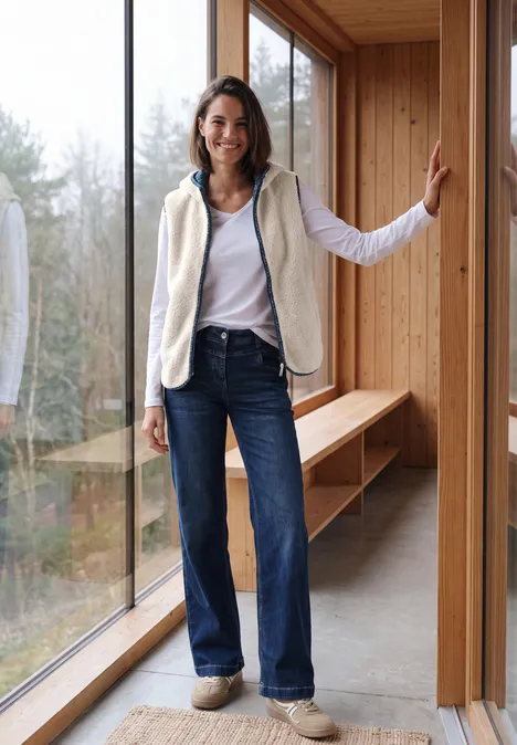 Eine Frau in weißem Top, Fleeceweste, weiten Jeans und Sneakern steht lächelnd an einem großen Fenster in einem Holzhaus.
