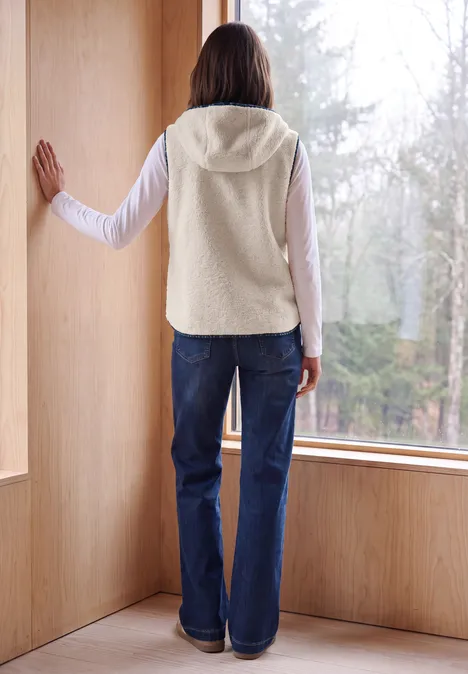 Person in Fleece-Weste und Jeans steht in einem Holzraum vor grossem Fenster und blickt in den Wald.