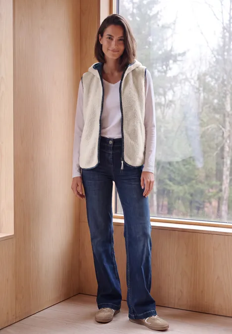 Eine Frau steht lässig in heller Fleecejacke und Jeans vor einem großen Fenster mit Blick auf den Wald.