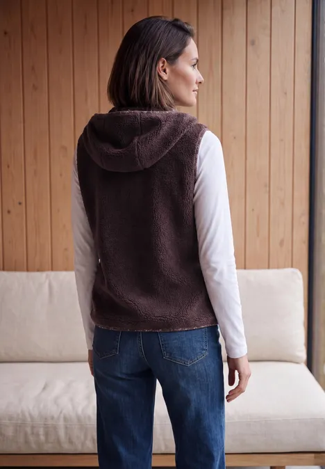 Rückenansicht einer Frau in brauner Fleece-Kapuzenweste, weißem Langarmshirt und Jeans vor Holzpaneel.