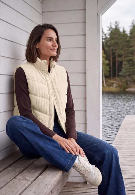 Eine Frau sitzt entspannt auf einer Terrasse am See, trägt Jeans, Pullover und Steppweste.