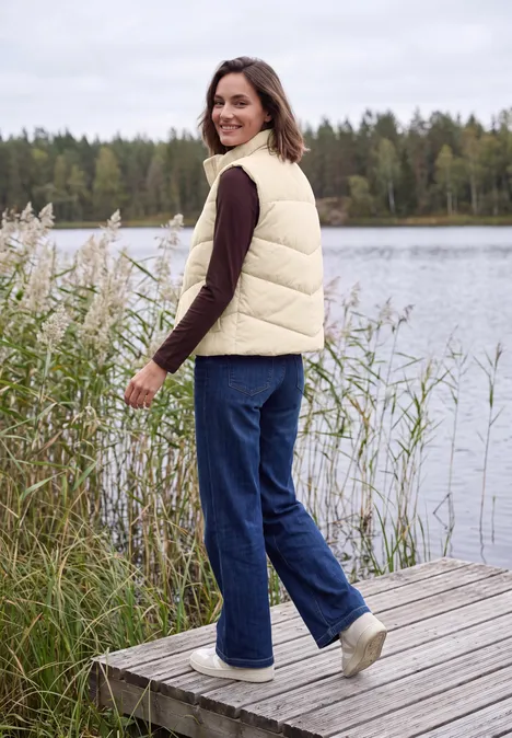 Frau im cremefarbenen Steppweste und Jeans steht auf einem Holzsteg am See, schaut lächelnd zurück.