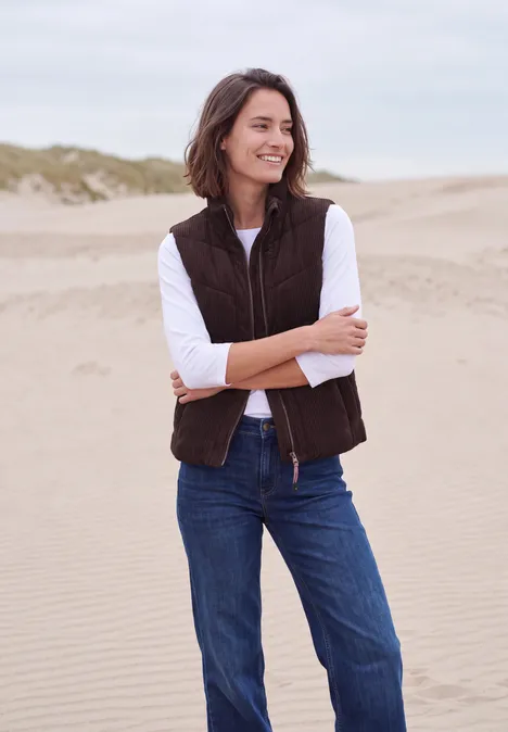 Een vrouw lacht op het strand met een bruin gilet, wit shirt en blauwe jeans voor zandduinen.