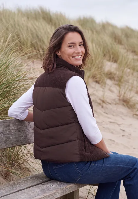 Glimlachende vrouw in bruine bodywarmer zit op een bankje in de duinen