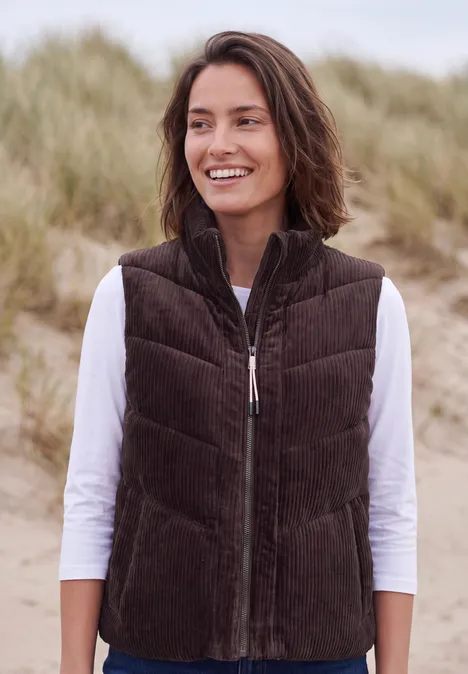 Een vrouw glimlacht bij zandduinen aan zee, ze draagt een bruine corduroy bodywarmer en een wit shirt met lange mouwen.
