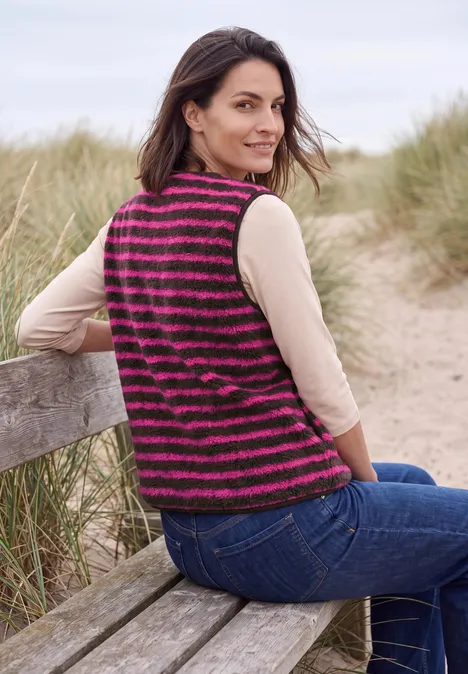 Frau im gestreiften pink-schwarzen Strickweste sitzt auf einer Bank am Strand und blickt lächelnd über die Schulter.