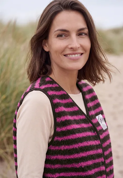 Eine lächelnde Frau trägt am Strand eine schwarz-pink gestreifte Strickweste und ein beiges Shirt.