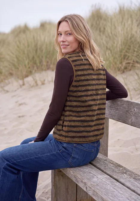 Teddy-Weste mit V-Neck und Streifen creamy mocha Eine Frau mit gestreiftem Strickweste sitzt auf einer Holzbank am Strand und lächelt.