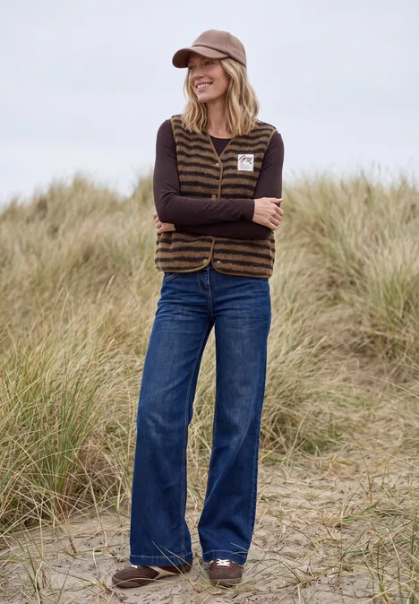 Teddy-Weste mit V-Neck und Streifen creamy mocha Frau mit Baseballcap, gestreiftem Teddyweste, schwarzem Langarmshirt und weit geschnittenen Jeans steht in Dünen.