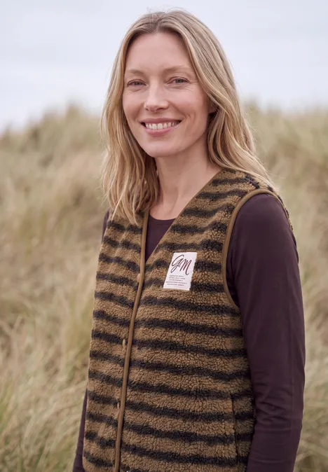 Teddy-Weste mit V-Neck und Streifen creamy mocha Eine blonde Frau mit gestreifter Weste steht lächelnd vor grasbewachsenen Dünen.