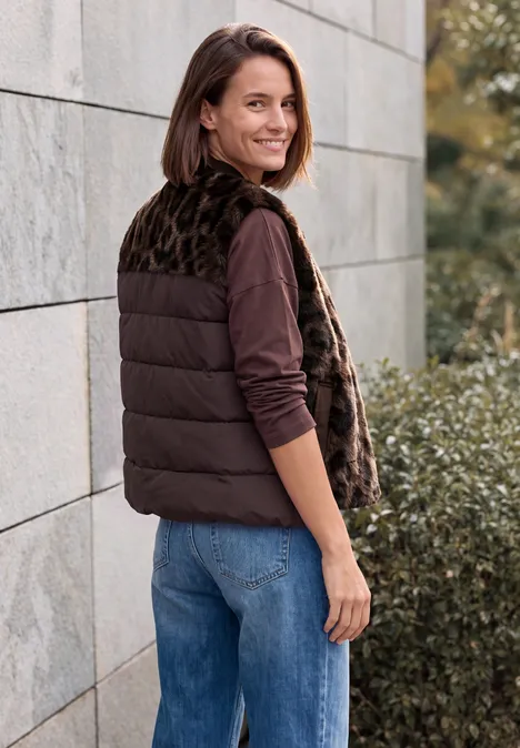 Teddyvest met gewatteerde details en patroon truffle brown Eine lächelnde Frau in brauner Steppweste, Jeans und langärmeligem Shirt steht seitlich vor Steinwand und Büschen.