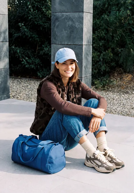 Teddyvest met gewatteerde details en patroon truffle brown Junge Frau mit Baseballcap und Freizeitkleidung sitzt lächelnd im Freien neben einer blauen Sporttasche.