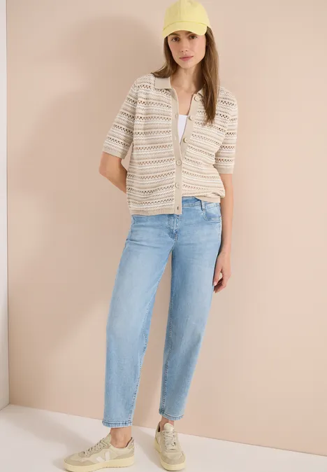 Frau trägt kurzärmliges gestreiftes Hemd, weiße Bluse, lockere blaue Jeans und bequeme Sneaker vor hellem Hintergrund.