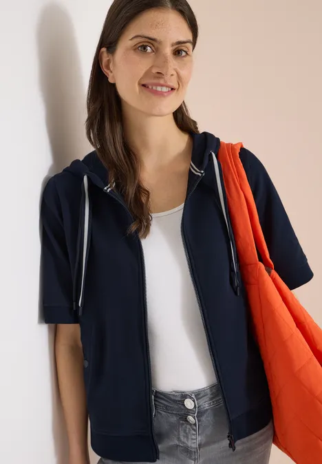 Kurzarm Sweatjacke mit Kapuze universal blue Person in dunkelblauer Kapuzenjacke über weißem Top trägt eine orangefarbene Tasche vor hellem Hintergrund.