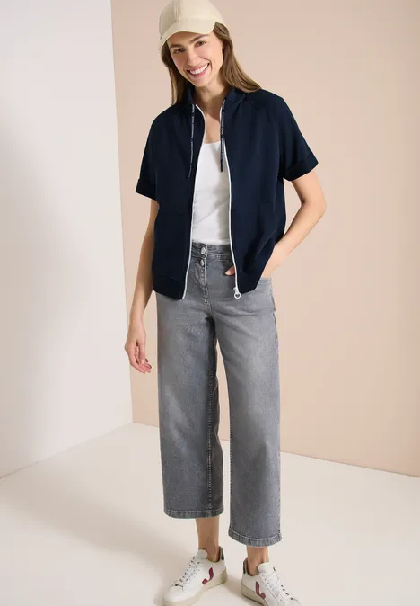 Kurzarm Sweatjacke mit Zipper universal blue Frau trägt weiße Bluse, offenes dunkelblaues Hemd, hellgraue, weite 7/8-Jeans und weiße Sneaker vor beigem Hintergrund.