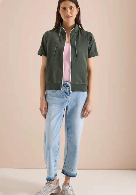 Kurzarm Sweatjacke mit Zipper khaki Modell in khakigrüner, kurzärmeliger Kapuzenjacke, rosa Top und hellblauen, umgeschlagenen Jeans.