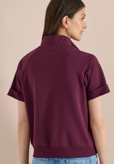 Kurzarm Sweatjacke mit Zipper sweet grape red Frau steht mit dem Rücken zur Kamera und trägt ein kurzes, auberginefarbenes Poloshirt mit umgekrempelten Ärmeln.