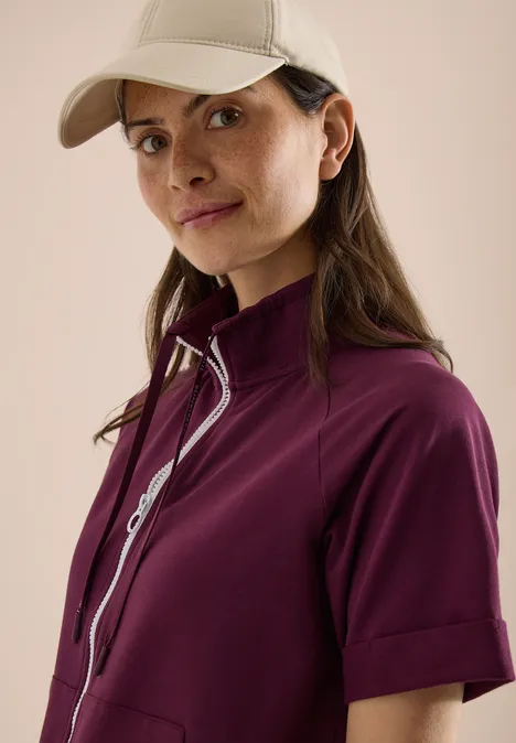 Kurzarm Sweatjacke mit Zipper sweet grape red Eine Person in einer lila Jacke und einer beigefarbenen Kappe vor hellem Hintergrund.