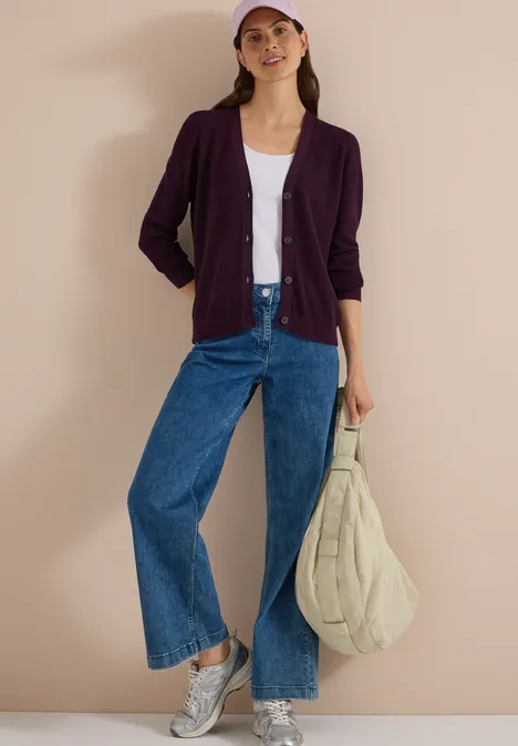 Frau in lila Cardigan, weißem Top, weiten Jeans, grauen Sneakern, rosa Cap und beiger Stepp-Tasche.