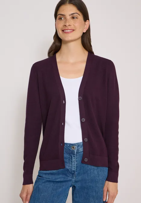 Frau trägt lila Strickjacke über weißem Top und blauen Jeans.