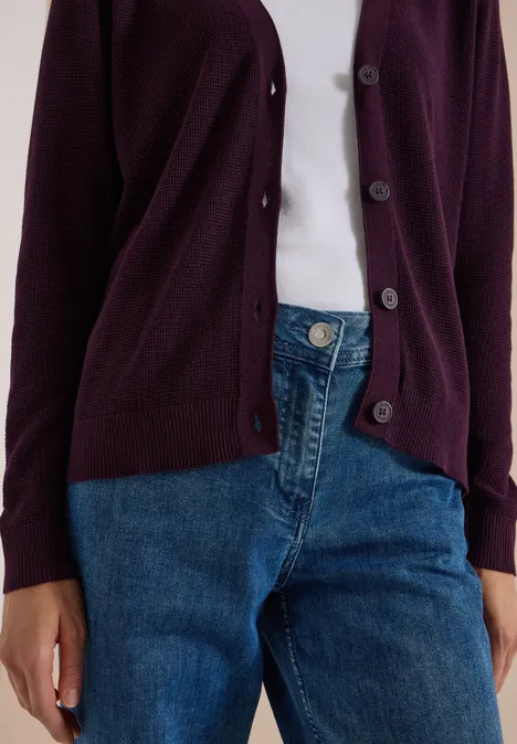 Person trägt lila Strickjacke über weißem Shirt und blaue Jeans.