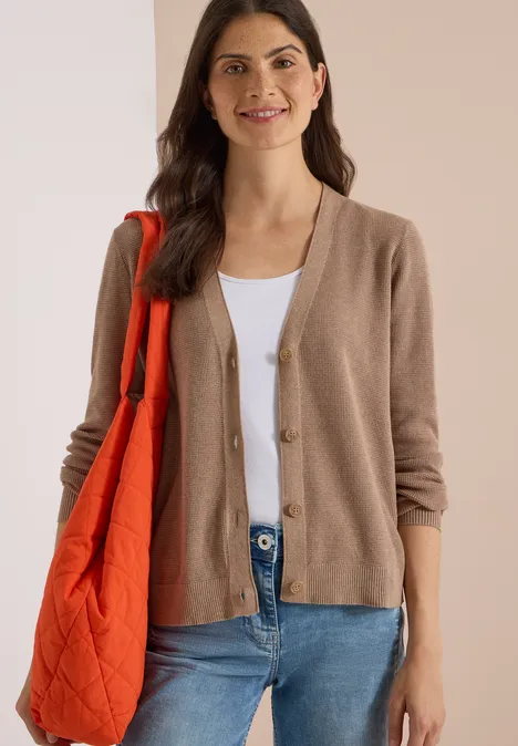 Braunhaarige Frau mit beigefarbener Strickjacke, weißem Top, blauen Jeans und oranger Umhängetasche lächelnd.