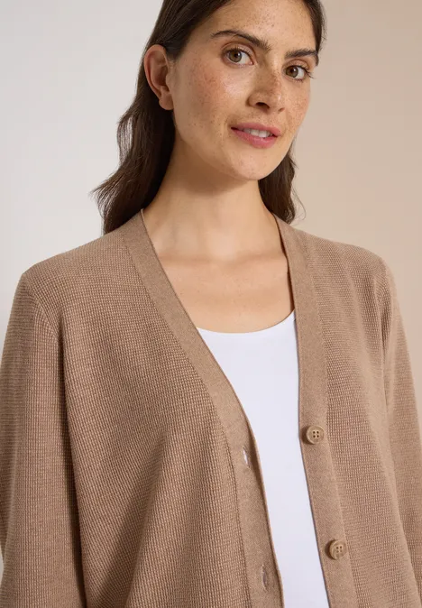 Eine Frau mit braunen Haaren trägt ein beiges Cardigan über einem weißen Top und schaut ruhig in die Kamera.