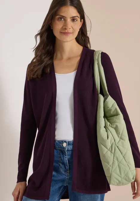 Frau in dunkelviolettem Cardigan, weißem Top, Jeans, mit großer, grüner Stepp-Tasche über der Schulter.