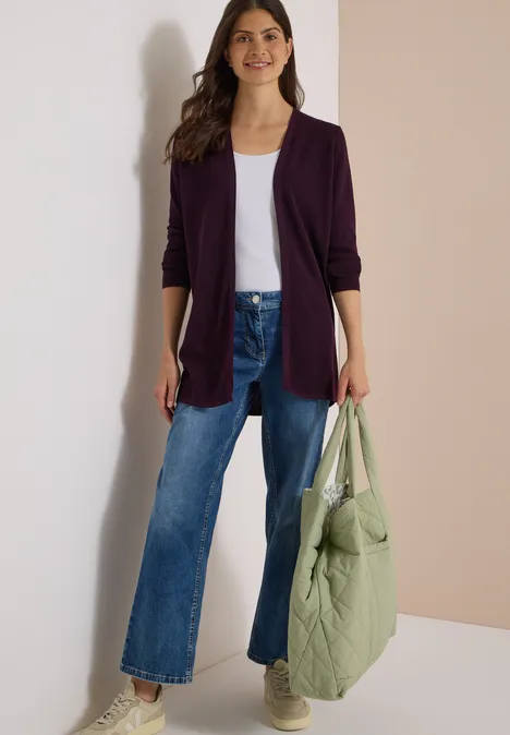 Frau in lila Cardigan, weißem Top, weiten Jeans und Sneakern hält einen hellgrünen Stepp-Beutel.