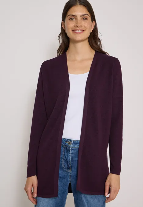 Frau in dunkelviolettem Cardigan, weißem Shirt und Jeans lächelt in die Kamera.