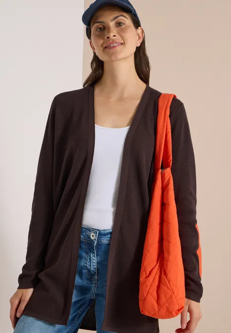 Frau in Jeans, weißem Top, dunkler Strickjacke, blauer Mütze und orangefarbener Tasche
