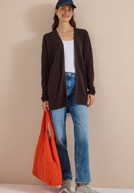 Frau trägt schwarzen Cardigan, weißes T-Shirt, blaue Jeans, rote Tasche und Mütze vor beigem Hintergrund.
