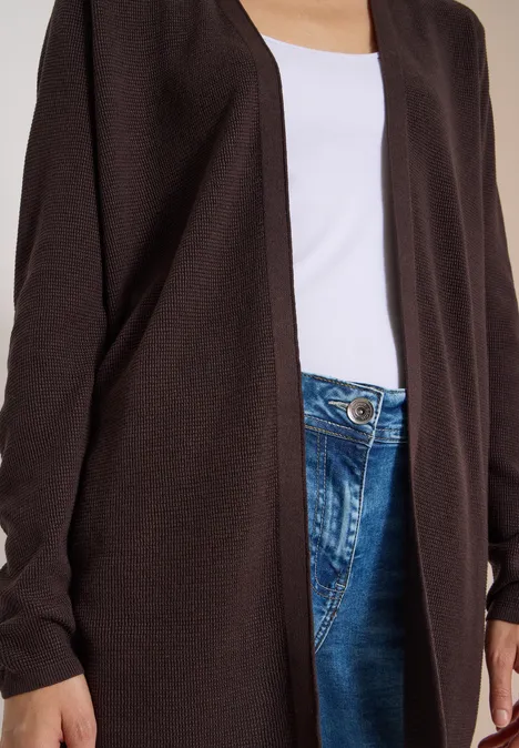 Person trägt offene dunkelbraune Strickjacke über weißem Top und blauen Jeans, nur Oberkörper sichtbar.