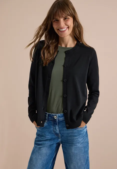 Een lachende vrouw draagt een groen shirt, een zwarte cardigan en spijkerbroek voor een neutrale achtergrond.