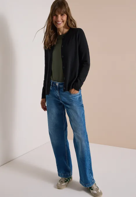 Een lachende vrouw in jeans, groen shirt en zwart vest.