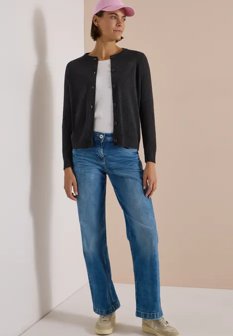 Eine Frau in schwarzer Strickjacke, weißem Top, blauen Jeans und rosa Basecap steht an einer Ecke.