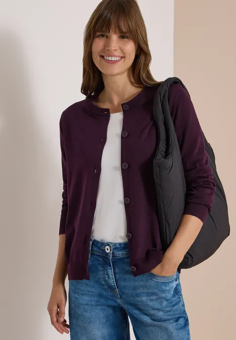 Cardigan mit Rundhals und Knopfleiste eggplant red Eine lächelnde Frau trägt eine lila Strickjacke, weißes Shirt, blaue Jeans und eine schwarze Schultertasche.