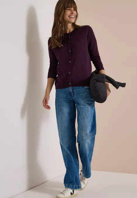 Cardigan mit Rundhals und Knopfleiste eggplant red Frau in lila Strickjacke und Jeans hält Jacke, geht lächelnd vor hellem Hintergrund.