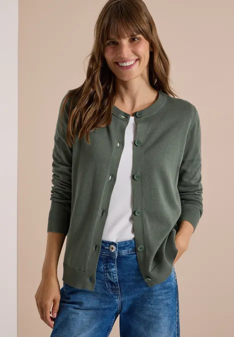 Glunderende vrouw in een groene cardigan, wit shirt en spijkerbroek voor neutrale achtergrond.