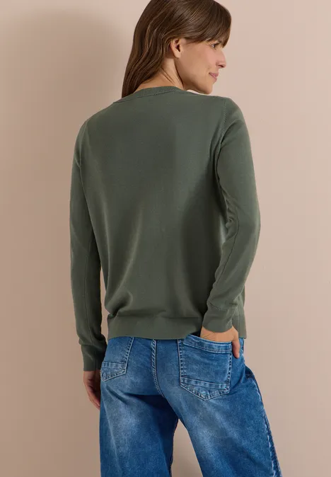 Een vrouw staat met haar rug naar de camera, in een groen trui en blauwe spijkerbroek voor een lichte achtergrond.