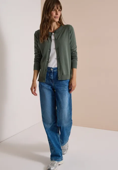 Een vrouw in een groen vest, wit T-shirt, blauwe wijde jeans en sneakers.