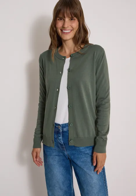 Vrouw lacht in een groen vest, wit shirt en blauwe jeans voor een witte muur.