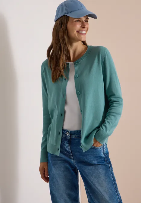 Een glimlachende vrouw draagt een groen vest, een witte T-shirt, een blauwe jeans en een lichtblauwe pet.
