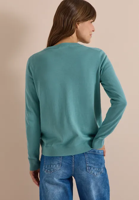 Vrouw van achteren in turquoise trui en spijkerbroek tegen neutrale achtergrond.