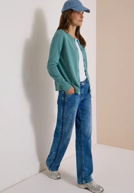 Vrouw tegen muur, groene cardigan, wit shirt, wijde jeans, sneakers en pet.