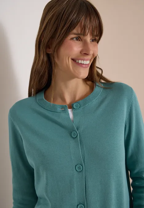 Vrouw lacht en kijkt naar rechts, draagt een groenblauwe gebreide cardigan.