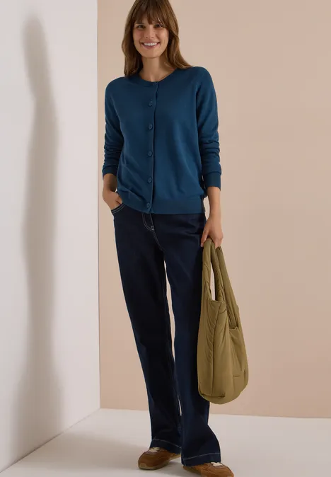 Cardigan mit Rundhals und Knopfleiste moonlight blue Frau in blauer Strickjacke und dunklen Jeans trägt eine grüne Tasche und lächelt vor schlichtem Hintergrund.