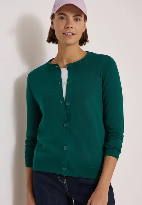 Frau trägt grüne Strickjacke, weißes T-Shirt, blaue Jeans und rosa Baseballcap vor weißem Hintergrund.
