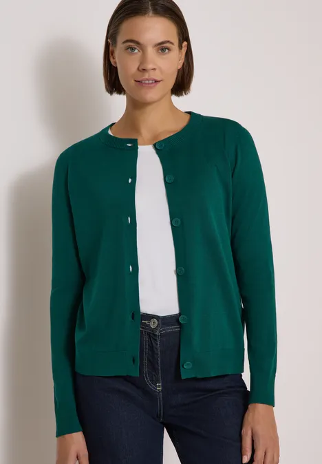 Frau trägt grünen Cardigan, weißes Shirt und Jeans vor hellem Hintergrund.